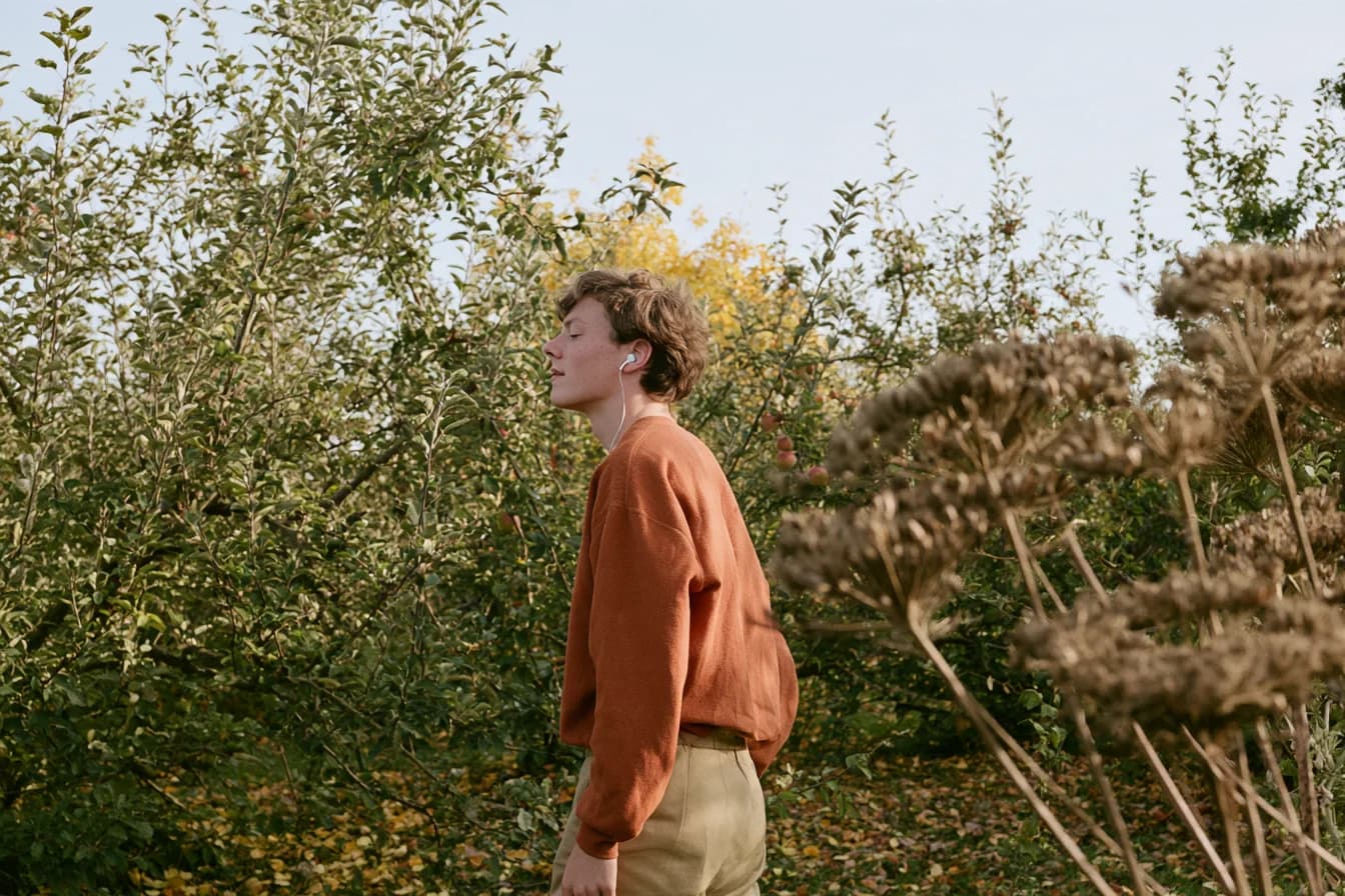 A visitor in an orange sweater listening to audio among orchard trees.